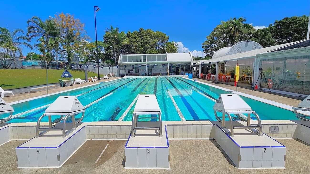 An outdoor swimming pool with multiple lanes, each marked by lane ropes. In the foreground, there are starting blocks numbered 2, 3, and 4, with "No Diving" signs. A swimmer is visible in the water. The background features a building with large windows and a curved roof, surrounded by trees and greenery. White chairs and a lifeguard stand are positioned along the poolside. The area is well-lit, suggesting a sunny day.