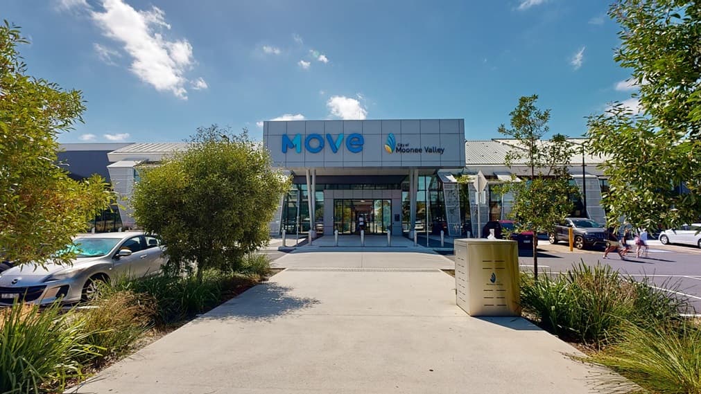 A two-storey building features a concrete and glass facade with an extended shade structure displaying a sign reading "Moonee Valley", flanked by larger "Move" letters. A large concrete post supports the shade, while metal mesh decorates the front. The building is topped with corrugated roofing.