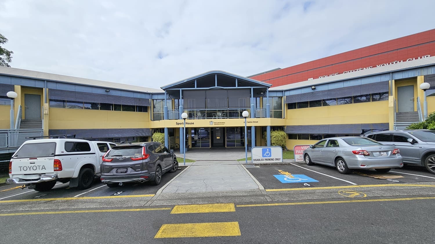 A two-storey building with a blue and yellow facade is in the centre, labelled "Sports House" with the address "150 Caxton Street." The foreground has a tiled walkway leading to the entrance, flanked by blue pillars. On the left, there are green bushes and a small bench. On the right, another bench and more greenery. The background includes large windows with blinds on both floors. The floor is tiled at the entrance and surrounded by a well-maintained grass lawn.