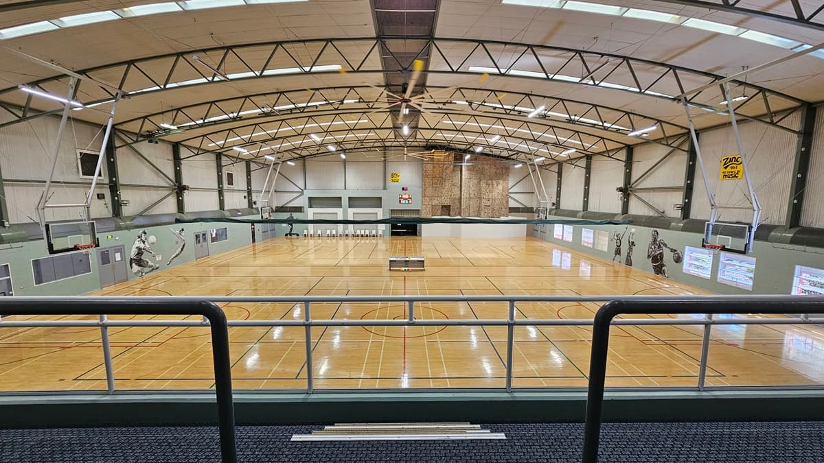 On the left are basketball hoops mounted on green walls with murals of players. In the middle is a large indoor basketball court with polished wooden flooring and various markings. The background features a climbing wall, a scoreboard, and rows of white chairs. On the right, there are more hoops and player murals. The ceiling is high with exposed beams, fluorescent lighting, skylights, and ventilation ducts. The foreground has a carpeted walkway with a metal handrail.