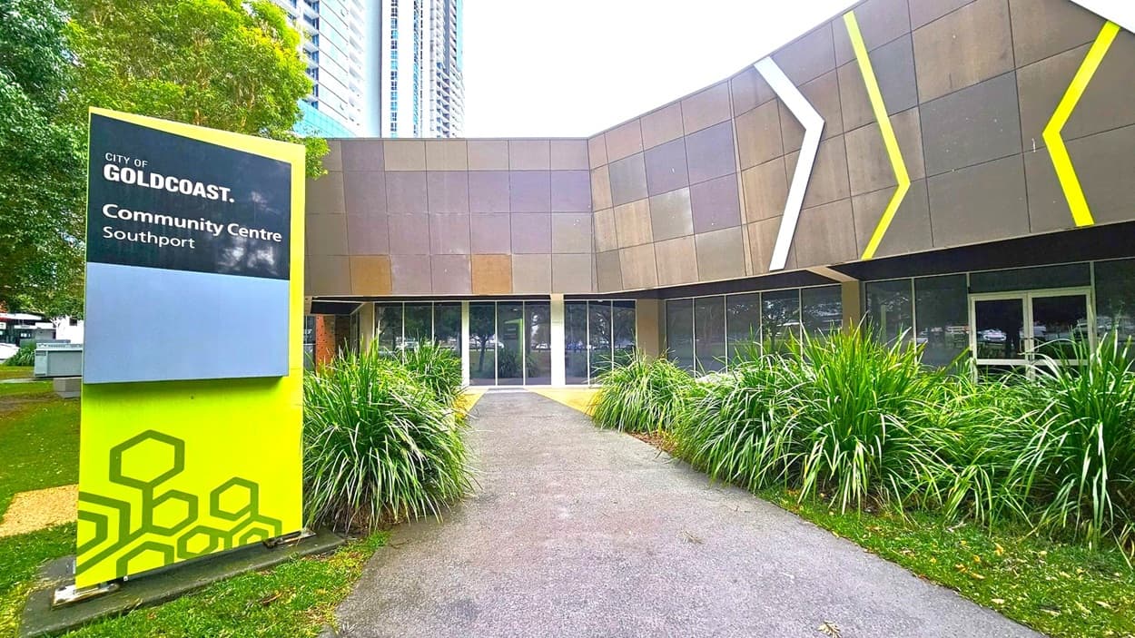 A modern building with a geometric facade featuring grey panels and diagonal white and yellow accents. In the foreground, a path leads to the entrance, flanked by lush green grass and plants. On the left, a sign reads "City of Gold Coast Community Centre Southport" in white text on a blue and green background. Trees with overhanging branches frame the scene, adding a natural element to the urban setting.
