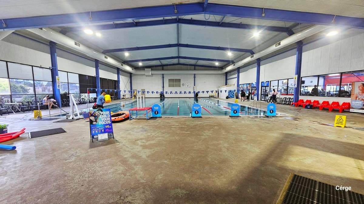25m, 5-lane swimming pool with ramp access and pool hoist in Portland Leisure and Aquatic Centre. On the right pool coping are two curved stainless steel grab rails near a yellow SLIPPERY WHEN WET sign. The pool lanes are separated by red, white, and blue floating rope lanes. Intersecting the pool are strings of triangular blue flags hanging above. The facility is a large steel structure with a white corrugated iron ceiling, blue steel beams, and large white ventilation ducts on both corners.