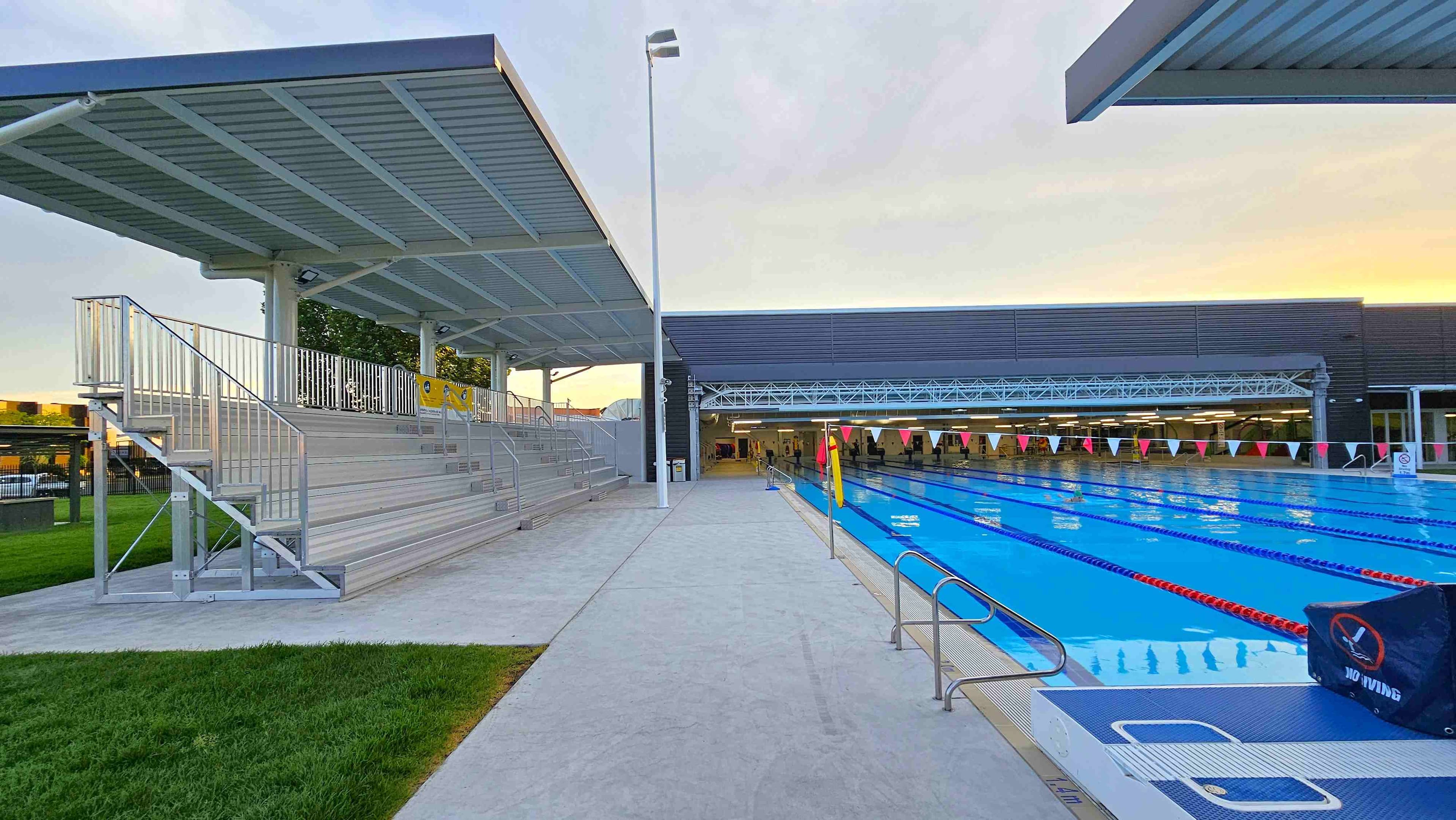 A modern building with a blue entrance and a sign reading "Inverell Aquatic Centre" in orange letters. The structure is surrounded by a well-maintained lawn and landscaped garden beds. A pathway leads from the street to the entrance. On the right, there is a red sign and some equipment. The foreground features a paved area and a road with a no-parking sign. The building has a flat roof and several small windows.