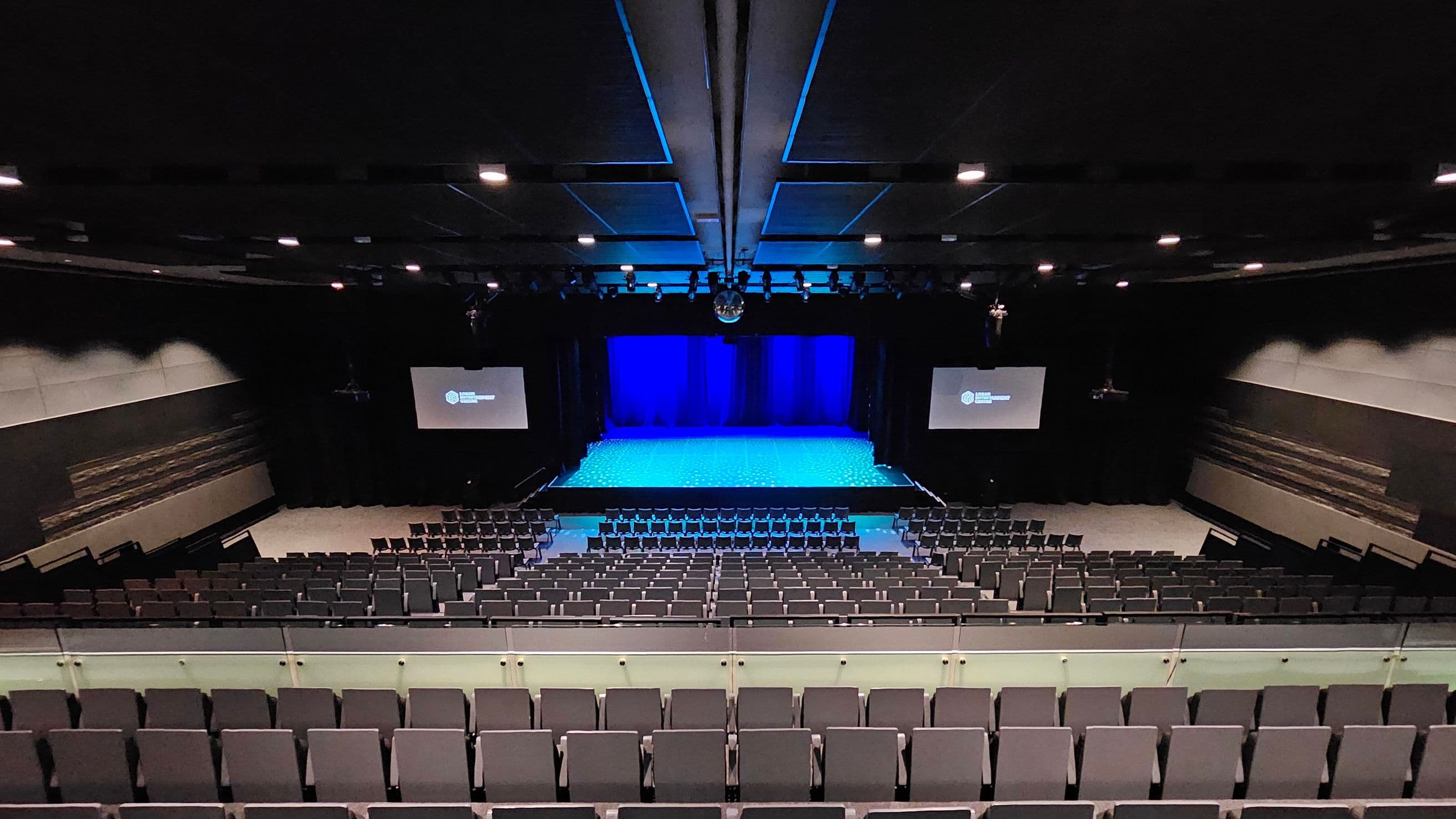 A large auditorium with tiered seating in the foreground, covered in dark fabric. The stage is in the centre, illuminated with blue lighting, and flanked by two large screens displaying text. The walls are lined with acoustic panels, and the ceiling features recessed lighting and sound equipment. The floor is carpeted in a dark colour, complementing the overall dimly lit atmosphere.