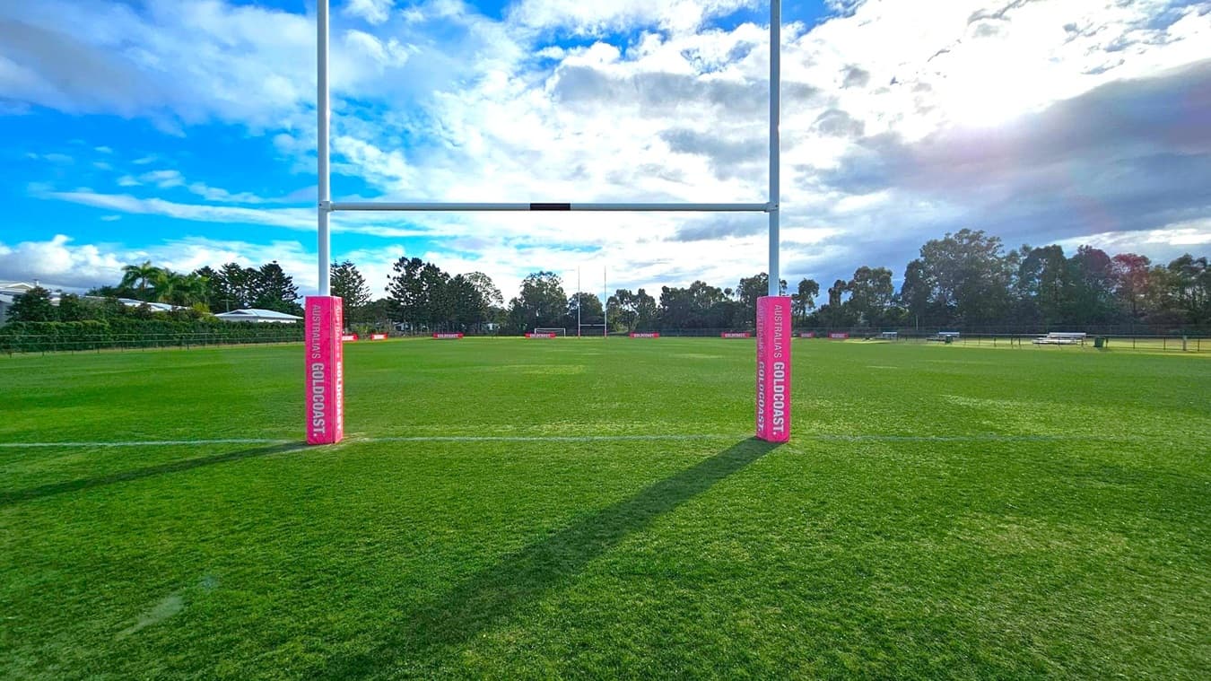 A rugby field with lush green grass. In the centre, there are rugby goalposts with a football goal behind them. Red advertising boards with "GOLDCOAST." are placed along the fence. Tall trees form a dense line in the background, providing a natural border. The sky is partly cloudy, casting a soft light over the scene.