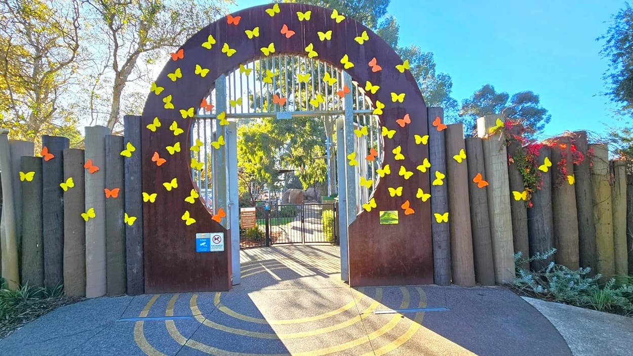 A decorative entrance gate with a circular arch adorned with yellow and orange butterfly cutouts. The gate is flanked by vertical wooden posts. Beyond the gate, a pathway leads into a garden area with trees and plants. The ground features a pattern of concentric circles in shades of grey and yellow. Trees with green foliage are visible in the background, and there are signs on the gate.