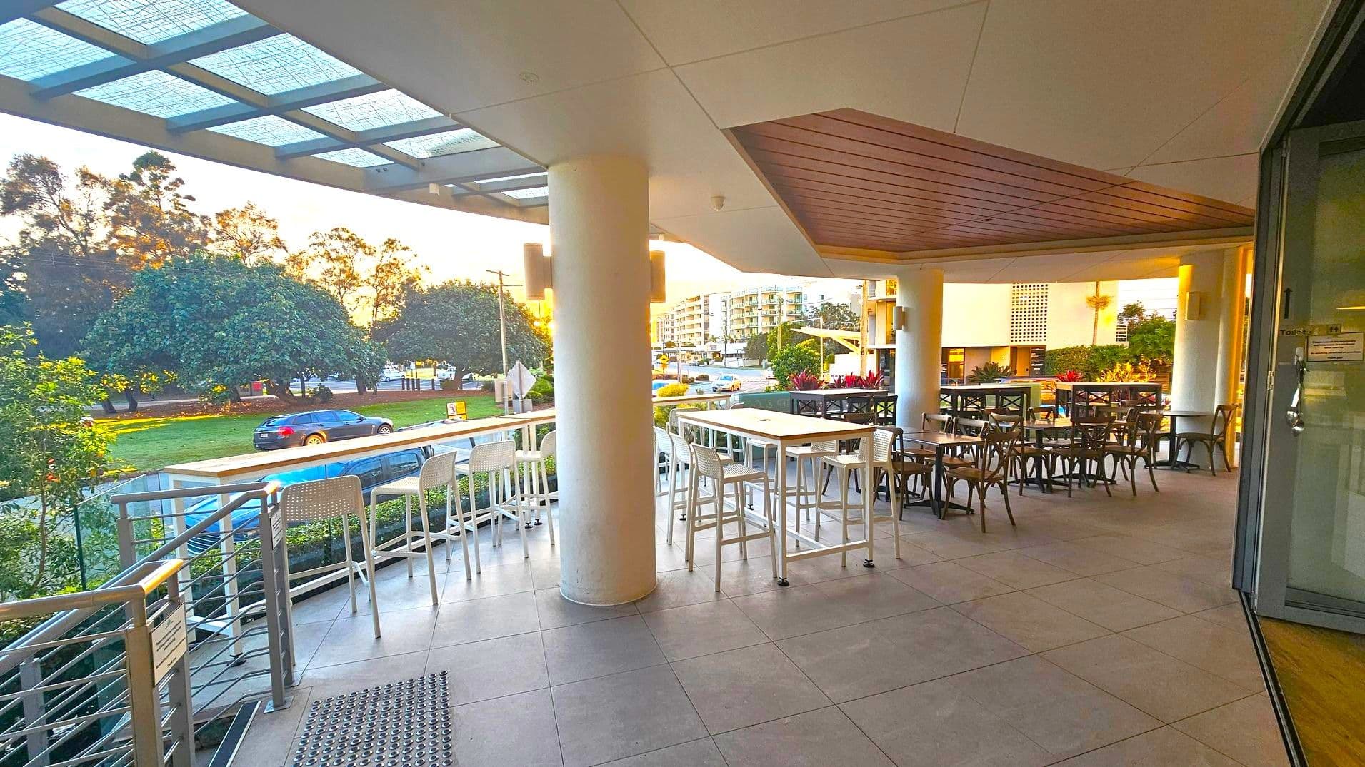 On the left, there are tall white bar stools lined up against a railing, with a view of trees and a road in the background. In the middle, a large white column supports the ceiling, and there are more tables and chairs. On the right, there are additional tables and chairs, with a glass door leading inside. The floor is tiled in grey, and the ceiling has a wooden panel section above the seating area.