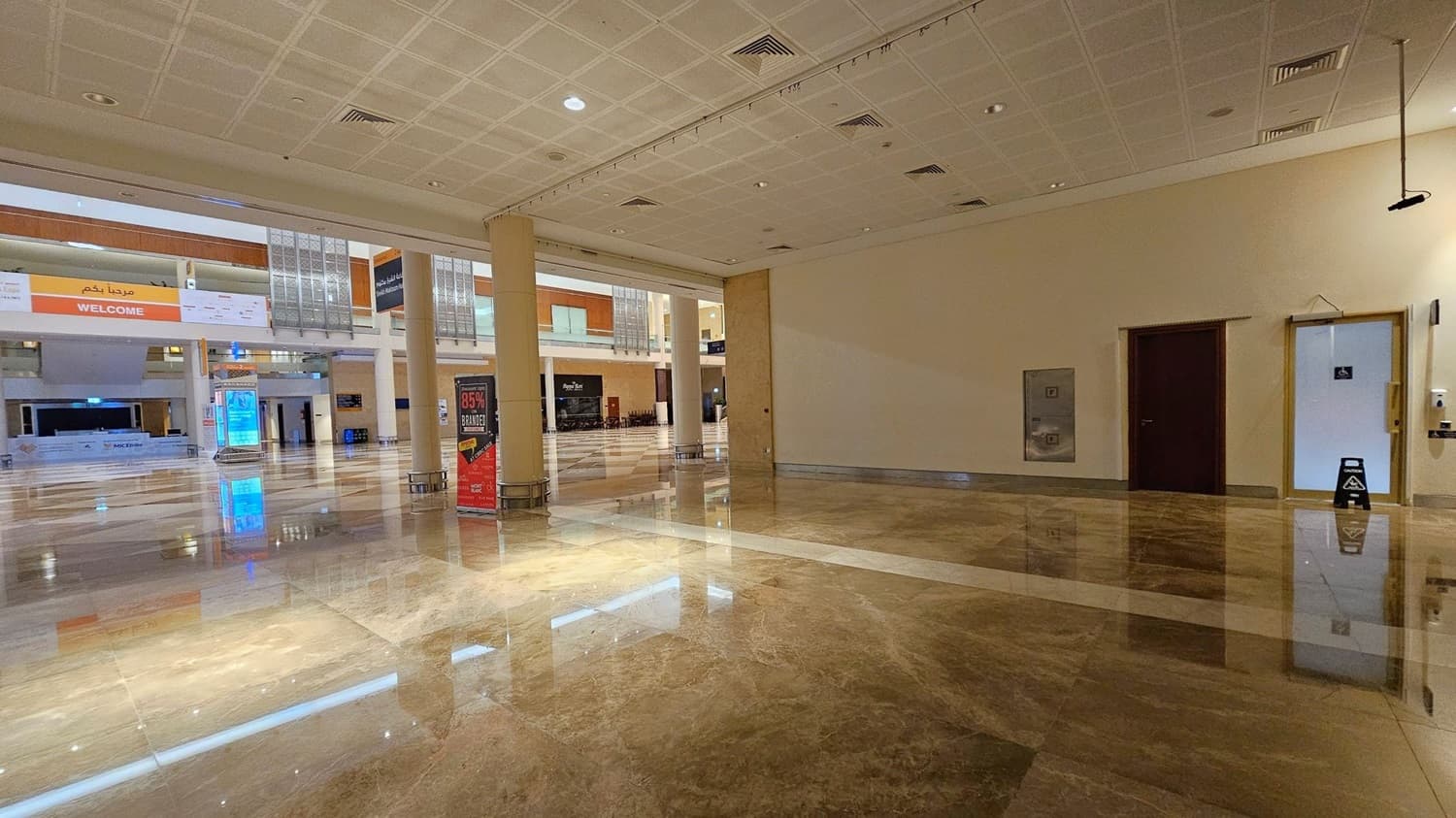 A spacious indoor area with a shiny marble floor. On the left, there's a door with a disability sign and a wall-mounted hand sanitiser dispenser. In the centre, a small white partitioned area is set up. To the right, a large open space with columns and a colourful sign advertising a sale. In the background, a second level with wooden panels and a "WELCOME" banner is visible. The ceiling is tiled with recessed lighting.