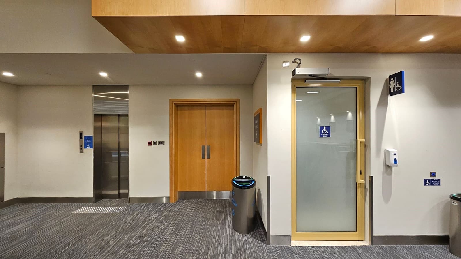 A hallway with a grey carpeted floor and a light-coloured ceiling with recessed lighting. On the left, there is a lift with a blue accessibility sign. In the centre, a wooden double door is flanked by a metal bin. On the right, a door with a glass panel has a sign for accessible toilets. Above it, a women's toilet sign is visible. A hand sanitiser dispenser is mounted on the wall next to the door.