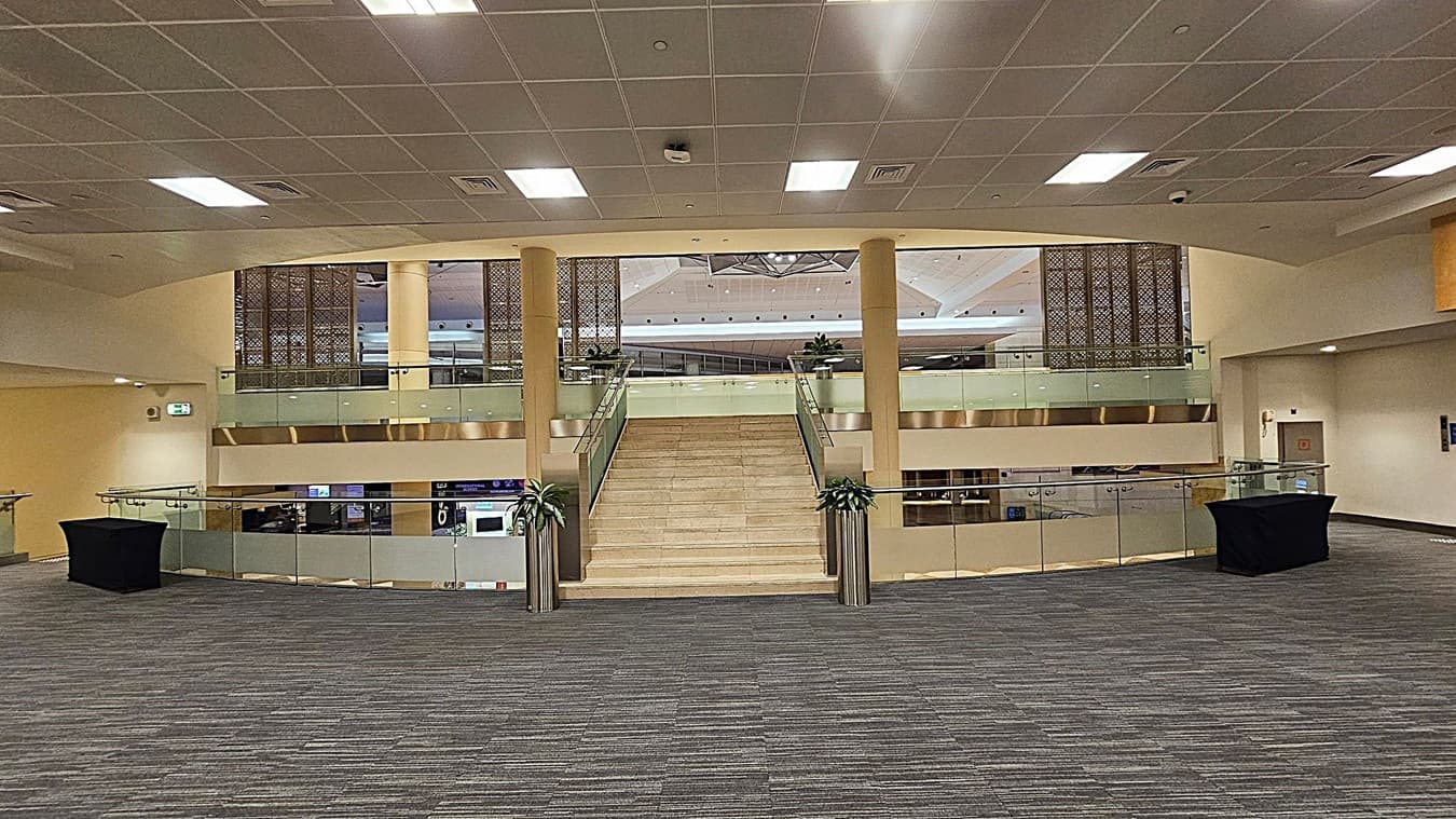 A spacious indoor area with a wide staircase in the centre leading to an upper level. The staircase is flanked by glass railings with metal accents. On either side of the staircase are large potted plants. The floor is carpeted in a grey and green pattern. The ceiling is fitted with square panels and recessed lighting. On the left and right, there are black covered tables. The background features decorative panels and a glimpse of a ceiling with geometric patterns.