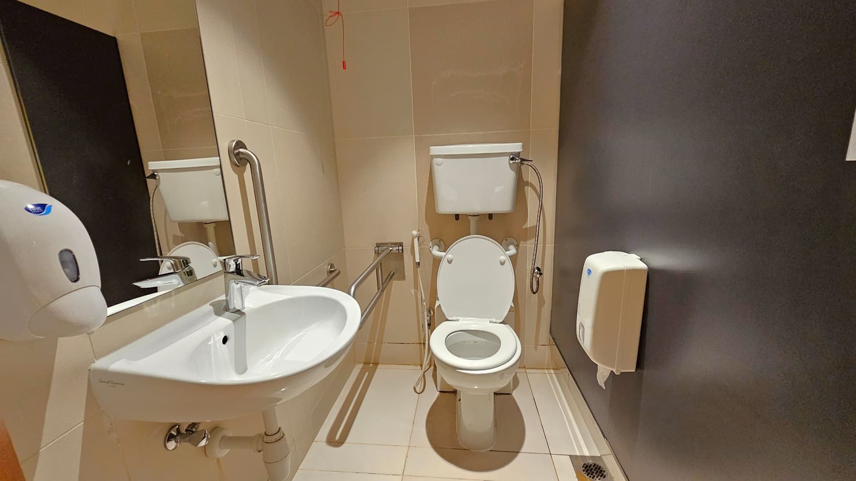 A bathroom with beige tiled walls and floor. On the left, a white sink with a chrome tap is mounted on the wall, accompanied by a soap dispenser and a large mirror. In the centre, a toilet with a white cistern and a handheld bidet spray is installed. Stainless steel grab bars are positioned around the toilet. On the right, a toilet paper dispenser is attached to a dark grey wall. A red emergency pull cord hangs from the ceiling.