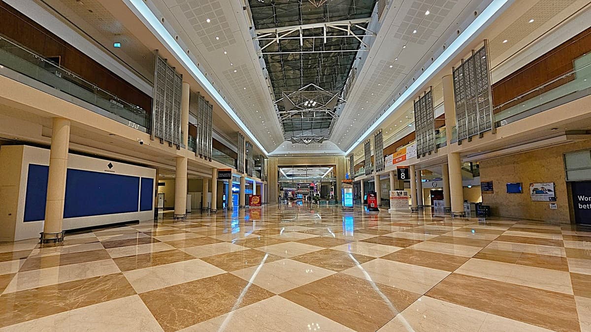 A spacious indoor area with a polished, chequered floor in shades of beige and brown. The ceiling is high with a geometric design and bright lighting. On both sides, there are two levels with glass railings and decorative panels. Various signs and digital displays are visible along the walls. In the background, there are more signs and a few people walking.