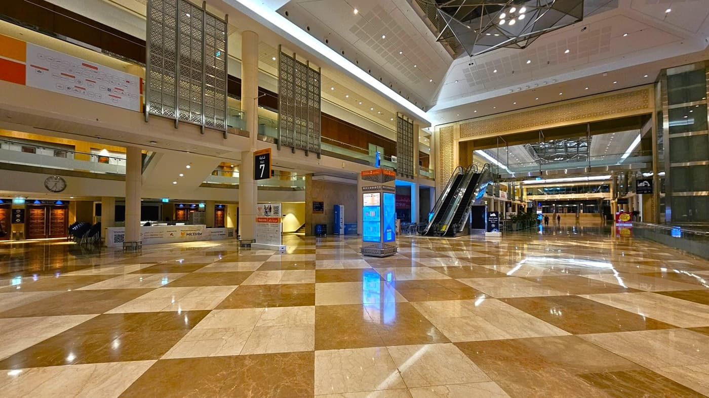 A spacious indoor area with a polished, chequered marble floor. On the left, there are information desks and a large wall clock. In the centre, a digital information kiosk stands near escalators leading to an upper level. The ceiling is high with modern lighting fixtures. The background features a large open space with more seating and signage. The area is well-lit and appears to be part of a public or commercial building.