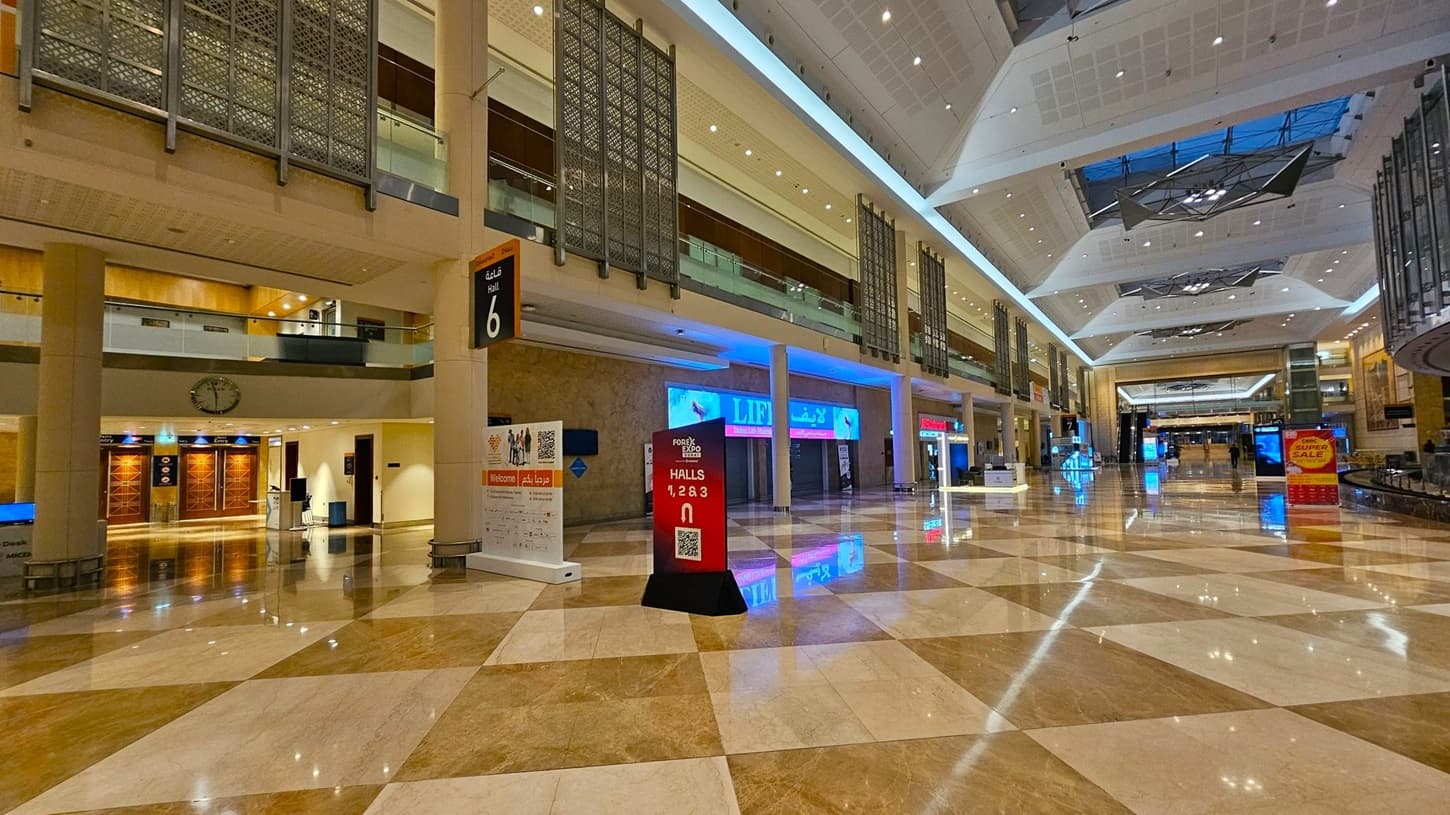 A spacious indoor hall with a polished, reflective marble floor in a checkered pattern of beige and brown. On the left, a large "WELCOME" sign in English and Arabic is displayed above a reception area. A digital kiosk with bright screens stands in the centre. On the right, a wide staircase leads to an upper level with glass railings. The ceiling is high with geometric designs and recessed lighting. Hall 8 is indicated by a sign hanging from the ceiling.