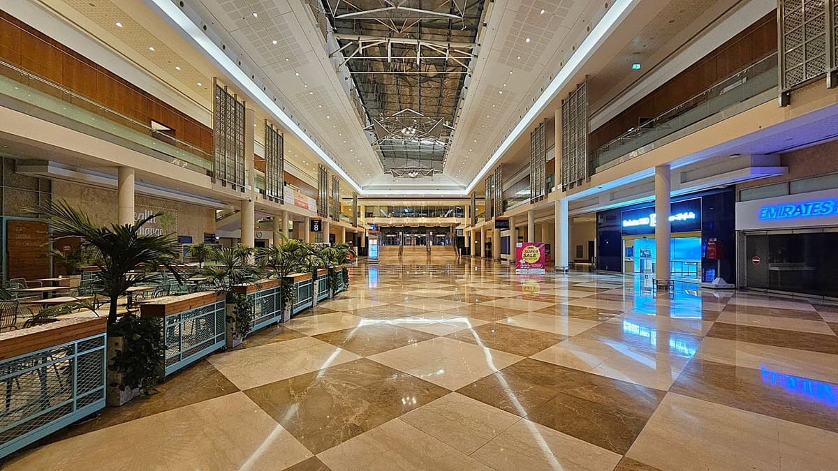 A spacious indoor area with a polished, chequered floor in shades of beige and brown. The ceiling is high with recessed lighting. In the centre, a large welcome banner is displayed above a reception desk. On the left, vending machines and a small seating area are visible. On the right, digital screens and signage for halls are present. The upper level features decorative panels and glass railings.