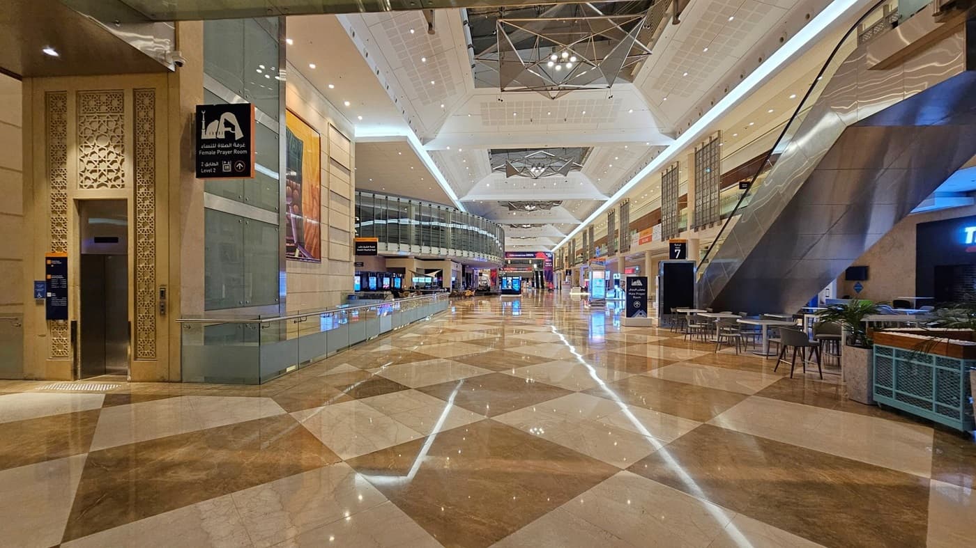A spacious indoor area with a polished, reflective floor featuring a geometric pattern. On the left, there's a sign for a female prayer room next to an intricately designed doorway. In the centre, a wide corridor extends with various shops and digital displays. On the right, an escalator leads to an upper level, with tables and chairs nearby. The ceiling is high with modern lighting fixtures and geometric designs.