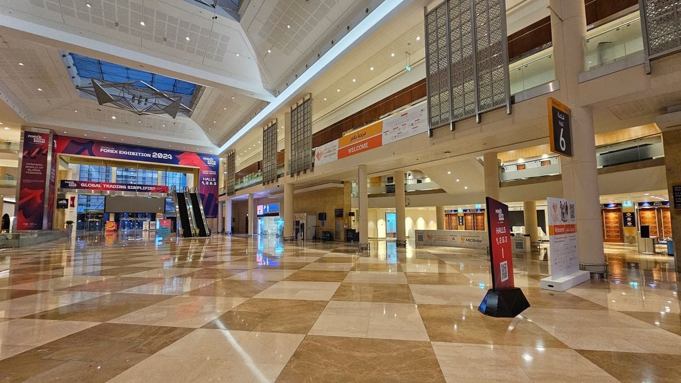A spacious exhibition hall with a polished, reflective marble floor in a checkered pattern. On the left, a large banner reads "Forex Exhibition 2024" above an entrance with glass doors. Two escalators are visible nearby. On the right, a sign indicates Hall 6, and there are various informational signs and banners. The ceiling is high with a geometric design and a skylight. The upper level features decorative panels and a "Welcome" banner.