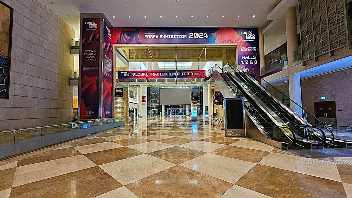 A spacious indoor area with a polished, chequered marble floor. On the left, there is a large wall with Forex Expo banners. In the centre, a wide entrance with a banner reading "Global Trading Simplified" leads to a hall. On the right, an escalator ascends to an upper level. The ceiling is high with recessed lighting, and the walls are adorned with additional banners and decorative panels.