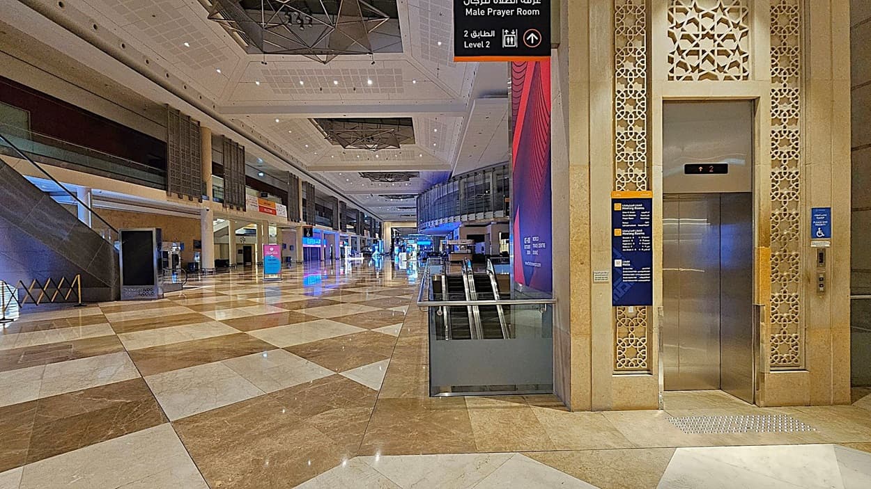 A spacious indoor area with a polished, patterned floor of brown and white tiles. On the right, there's an elevator with intricate decorative panels and signage indicating directions. In the centre, an escalator descends. The left side features a staircase and various signs. The ceiling is high with geometric designs and lighting fixtures. The background shows a long corridor with illuminated signs and displays.