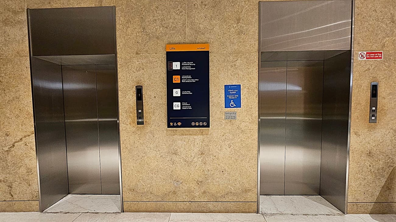 Two stainless steel lift doors are set into a beige marble wall. In the centre, a sign displays floor information in multiple languages. To the right of the sign, a blue notice indicates priority for wheelchair users. Each lift has a control panel with buttons and floor indicators. The floor is light-coloured stone, and the ceiling is not visible.