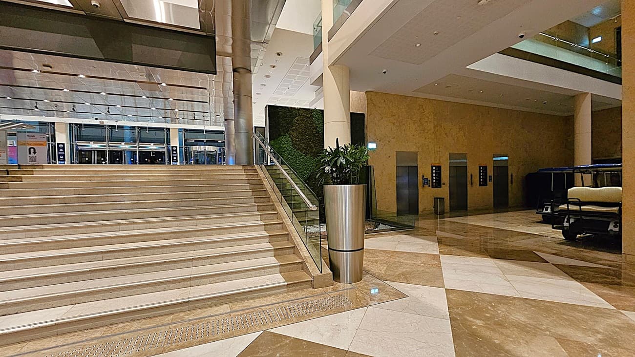 A spacious indoor area with a wide staircase on the left, featuring glass railings. In the centre, a large metal planter with green plants stands next to the stairs. The background shows a wall with a green vertical garden. On the right, there are several lifts with illuminated indicators and a small vehicle parked nearby. The floor is polished with a geometric pattern of light and dark tiles, and the ceiling is fitted with recessed lighting.