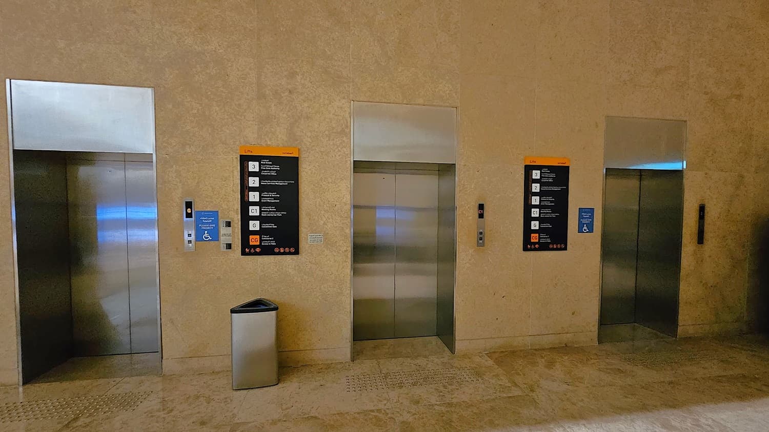 Three lift doors are set into a beige stone wall. Each lift has a metallic finish. On the left, a blue sign indicates accessibility, and a control panel is visible. In the centre, a black and orange directory lists floor numbers and destinations. A silver bin is placed on the floor in front of the middle lift. The floor is made of polished stone tiles. The ceiling is not visible.