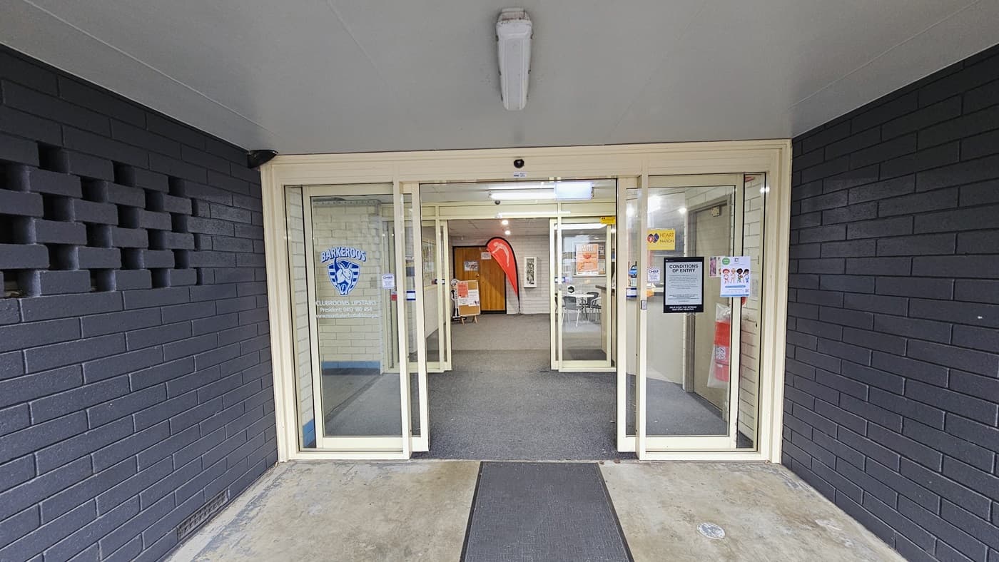 The entrance features automatic double glass doors surrounded by two dark blue brick walls. Posters and stickers with reminders adorn the glass doors. A non-slippery mat is placed on the doorstep. The ceiling is painted white and has fluorescent lights. Inside the building, chairs and banners are visible.