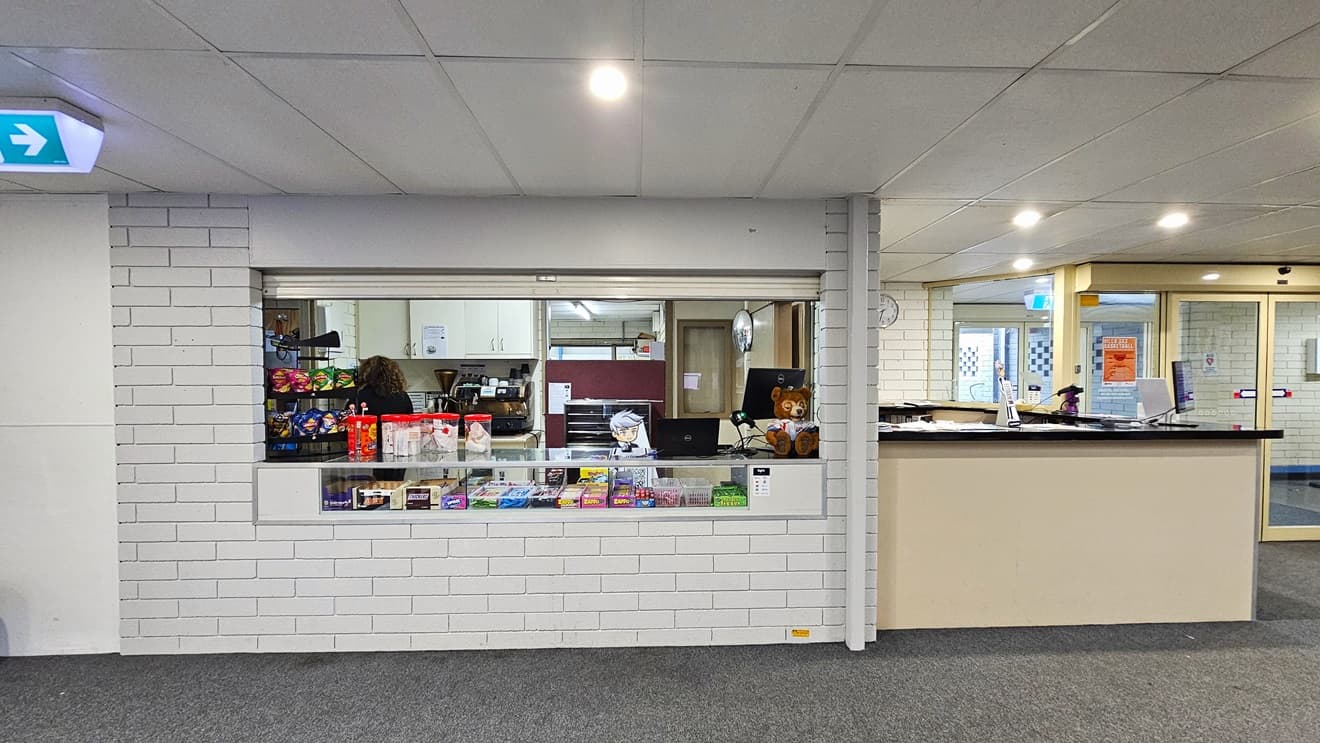 On the right is a white reception desk and an automatic glass door. In the middle is the cafe offering hot and cold drinks, desserts, and treats arranged on the display rack and on the counter. A computer, a POS machine, and a brown teddy bear are placed on the corner of the counter. The wall has a white brick design, the ceiling has a white porous design, and the floor is grey. A sign showing direction is placed on the ceiling.