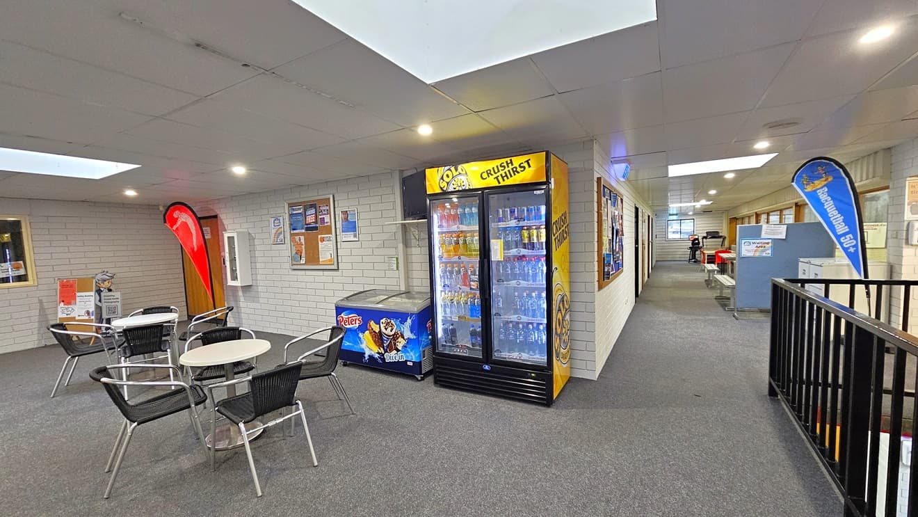 To the left, white tables and black chairs are arranged. A fridge stands in the corner. A corkboard with posters is on the wall, alongside safety reminders and a fire extinguisher. A door, a red flag, a display box, and a flatscreen TV are on the left. The lobby on the right leads to other areas, with banners, posters, and a metal railing visible.