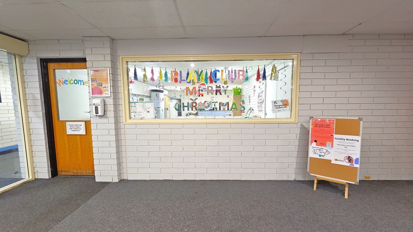 The Creche entrance, located to the left beside the glass door, features a wooden door with a welcoming sticker and reminder. A large window adorned with "Playclub" stickers allows a view inside, embellished with Christmas decorations. A framed poster and hand sanitizer dispenser adorn the white brick-designed wall. A freestanding board with additional reminders is positioned on the right of the window. The ceiling and floor maintain a neutral white and grey palette, respectively.