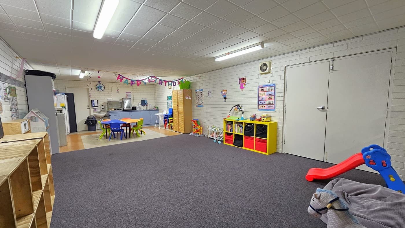 Inside the Creche, empty wooden display cabinets line the left side against the brick wall. Moving further, the kitchen area features a fridge on the left, a rubbish bin, and various kitchen accessories, including a sink. Brightly colored plastic chairs and tables are positioned in the middle. On the right, another display rack showcases toys. Toys are spread across the carpeted area, including a red-blue slide, a bean bag, and a horse ride. Colorful posters and banners are fixed on the walls.