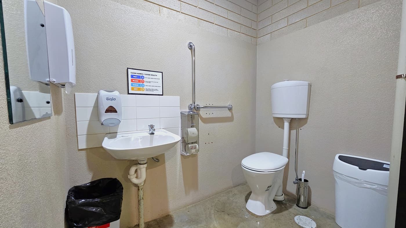 Inside the bathroom is a toilet bowl positioned in the left corner with handrails. On the left wall, a wash basin is mounted, accompanied by a soap dispenser, a tissue dispenser, and a mirror. To the right, a white rubbish bin is placed. The white wall features a brick accent design, and the floor has a marble design. Another rubbish bin is positioned below the wash basin, and a reminder poster is affixed to the left wall.