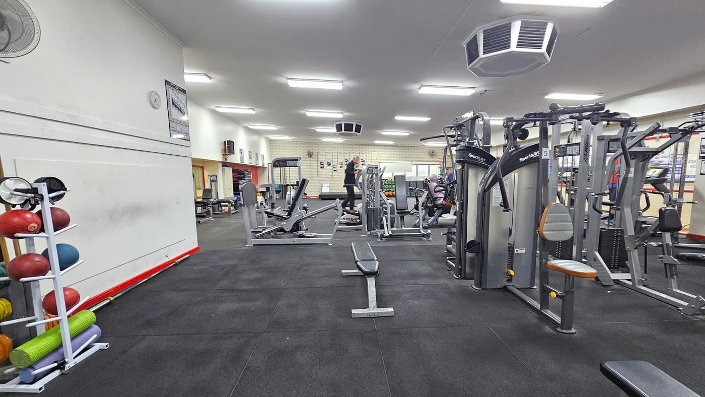 To the left is a display rack featuring medicine balls, weights, and rubber mats. A wall fan, clock, and poster adorn the top wall. On the right, various machines such as bench machines, Lat pull-ups, and treadmills are present. Two people are using the equipment. The floor is rubber tiles, and the white ceiling has rows of fluorescent lights with an octagon-shaped air conditioning unit mounted.
