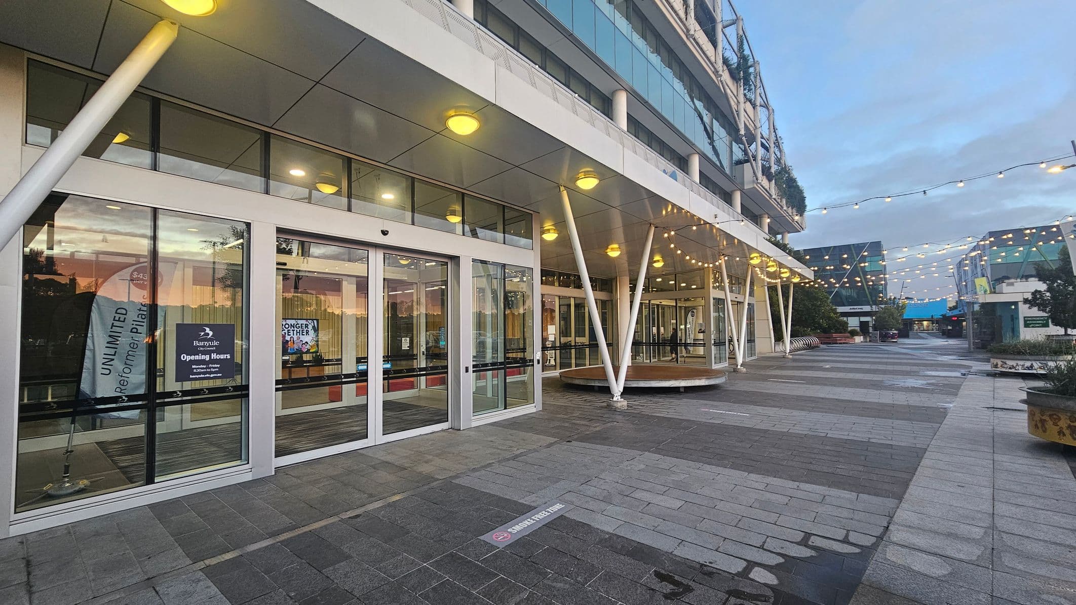 A modern building entrance with large glass doors on the left, displaying signs and banners. The ceiling features round lights, and the floor is paved with grey tiles. White structural beams support the overhang. To the right, a pedestrian walkway is adorned with string lights overhead. The area is open, with planters and benches along the path. The background shows more buildings with glass facades.