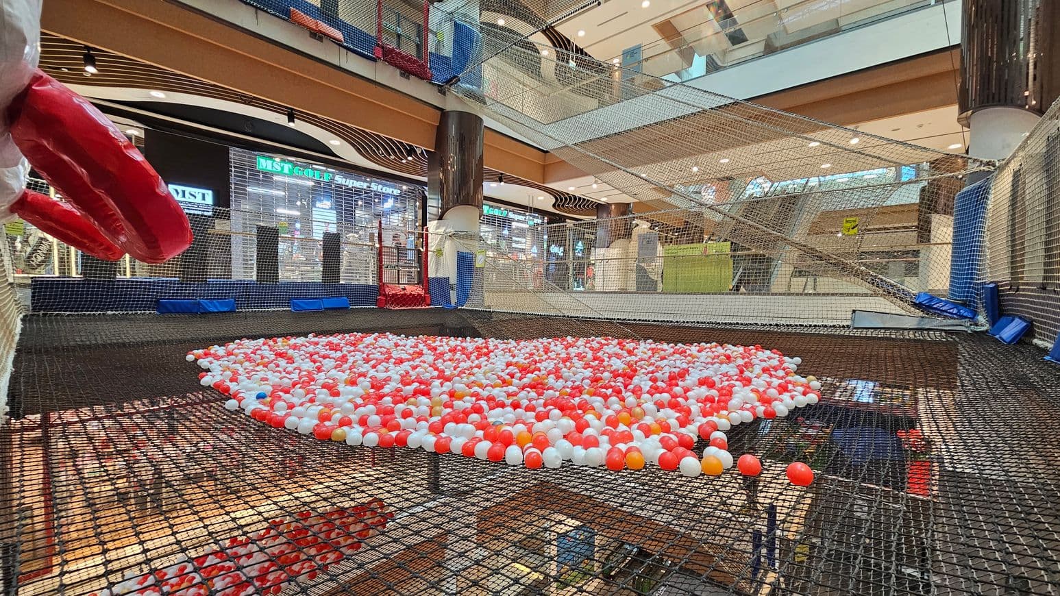 A shopping centre interior with a large netted play area in the foreground, filled with red, white, and orange plastic balls. On the left, a large red inflatable object is visible. The background features a store with a sign reading "MST GOLF Super Store" and other shops. The ceiling is high with multiple levels and glass railings, and the floor is covered with netting. The area is well-lit with overhead lights.