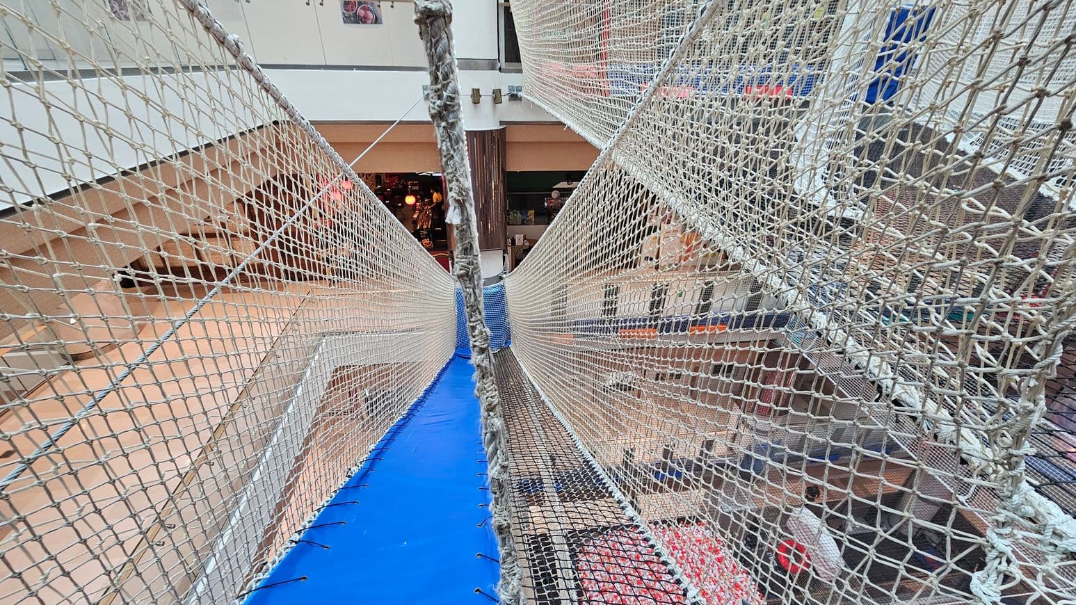 A suspended net bridge with blue padding on the floor stretches across a multi-level indoor space. The netting is tightly woven, forming walls on either side. Below, various floors of a shopping centre are visible, with shops and people. The background shows a glass railing and a column, with some lights and displays from the shops. The ceiling is not visible.