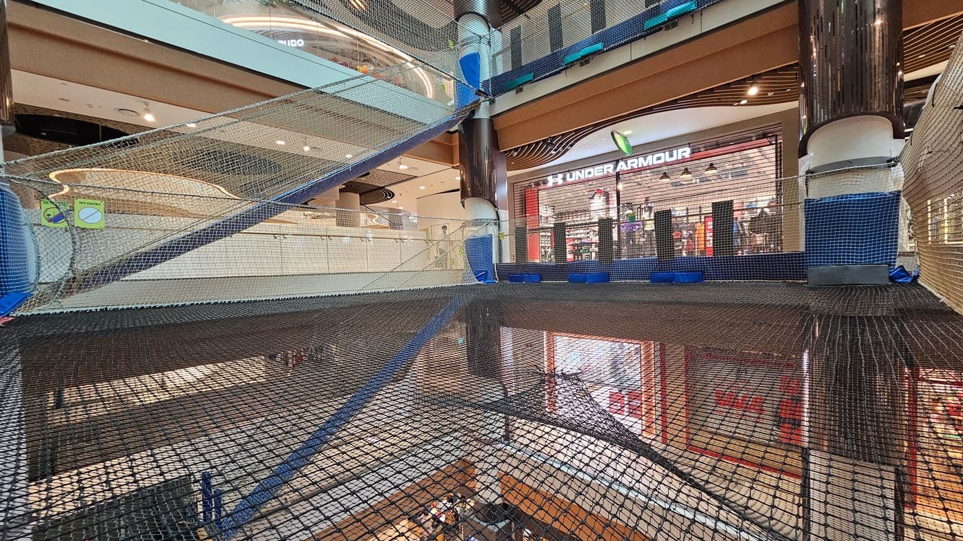 A shopping centre interior with multiple levels. In the foreground, there is a large net structure spanning across the space, supported by blue and black padding. On the right, a shop with a visible "Under Armour" sign. The background shows another level with a curved ceiling design and various lights. The floor is tiled, and there are signs indicating "No Entry" on the netting.