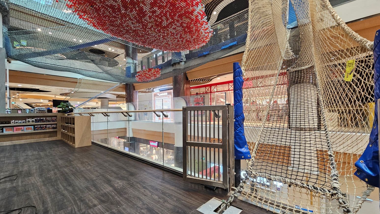 A shopping centre interior with a dark wooden floor. On the right, a netted play area with blue padding is suspended, leading upwards. In the middle, a glass railing overlooks the lower level, where shop signs like Levi's are visible. On the left, wooden shelves display various items. Above, colourful netting in red and blue is suspended from the ceiling, which features spotlights and wooden beams.