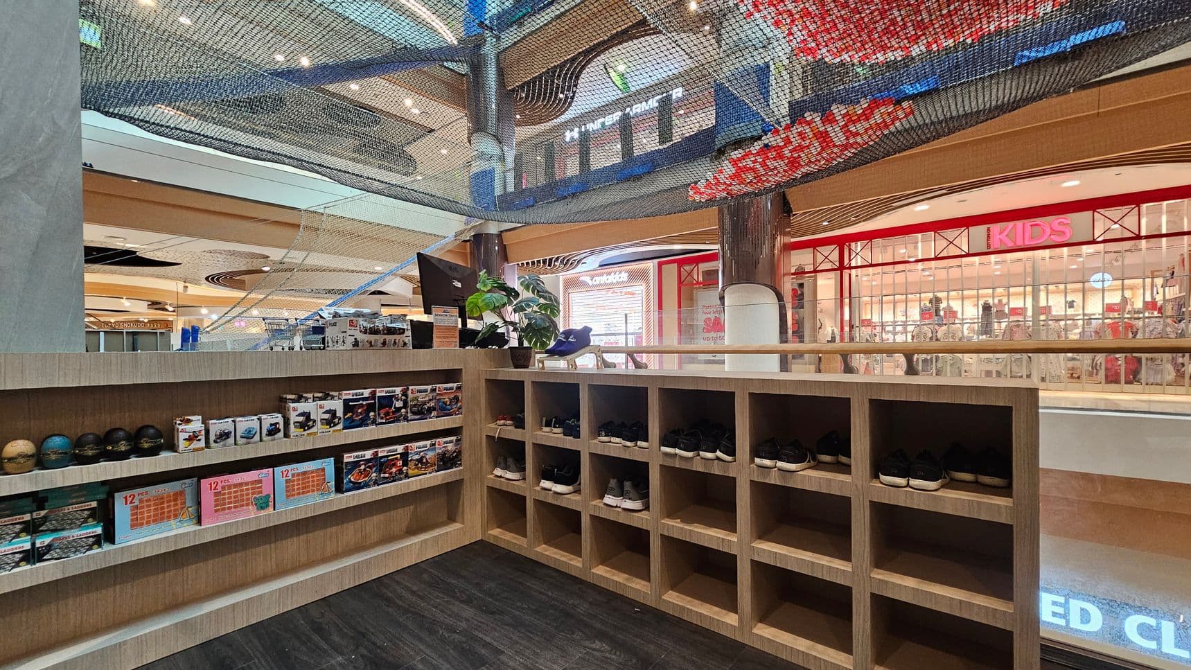 On the left, there are shelves displaying various boxed toys and small balls. In the middle, a wooden counter holds a plant and a pair of shoes. On the right, there are cubbyholes with more shoes. The background features a shop with a red sign reading "KIDS" and a glass storefront. Above, a netted structure is visible, and the ceiling has circular light fixtures. The floor is dark and smooth.