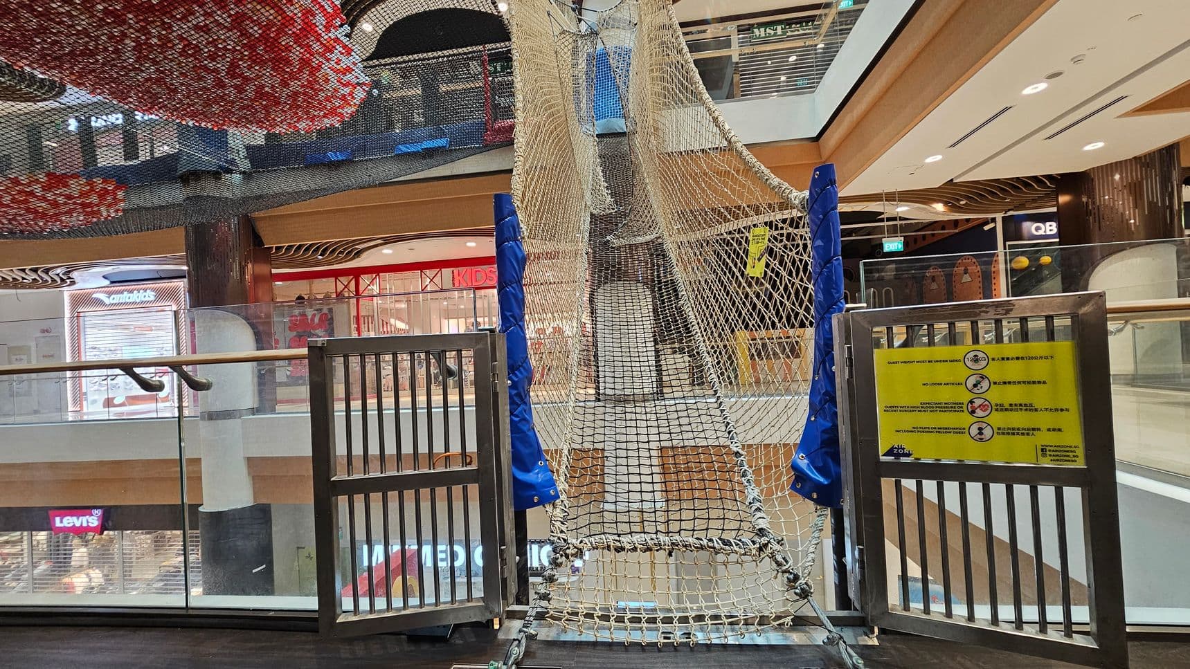 A shopping centre interior with a netted climbing structure in the foreground, supported by blue padded poles. A metal gate with a yellow sign displaying safety instructions is attached to the structure. In the background, there are various shopfronts, including Levi's and a children's store. The ceiling features decorative elements with red and black patterns. The floor is dark, and glass railings are visible on the left side.