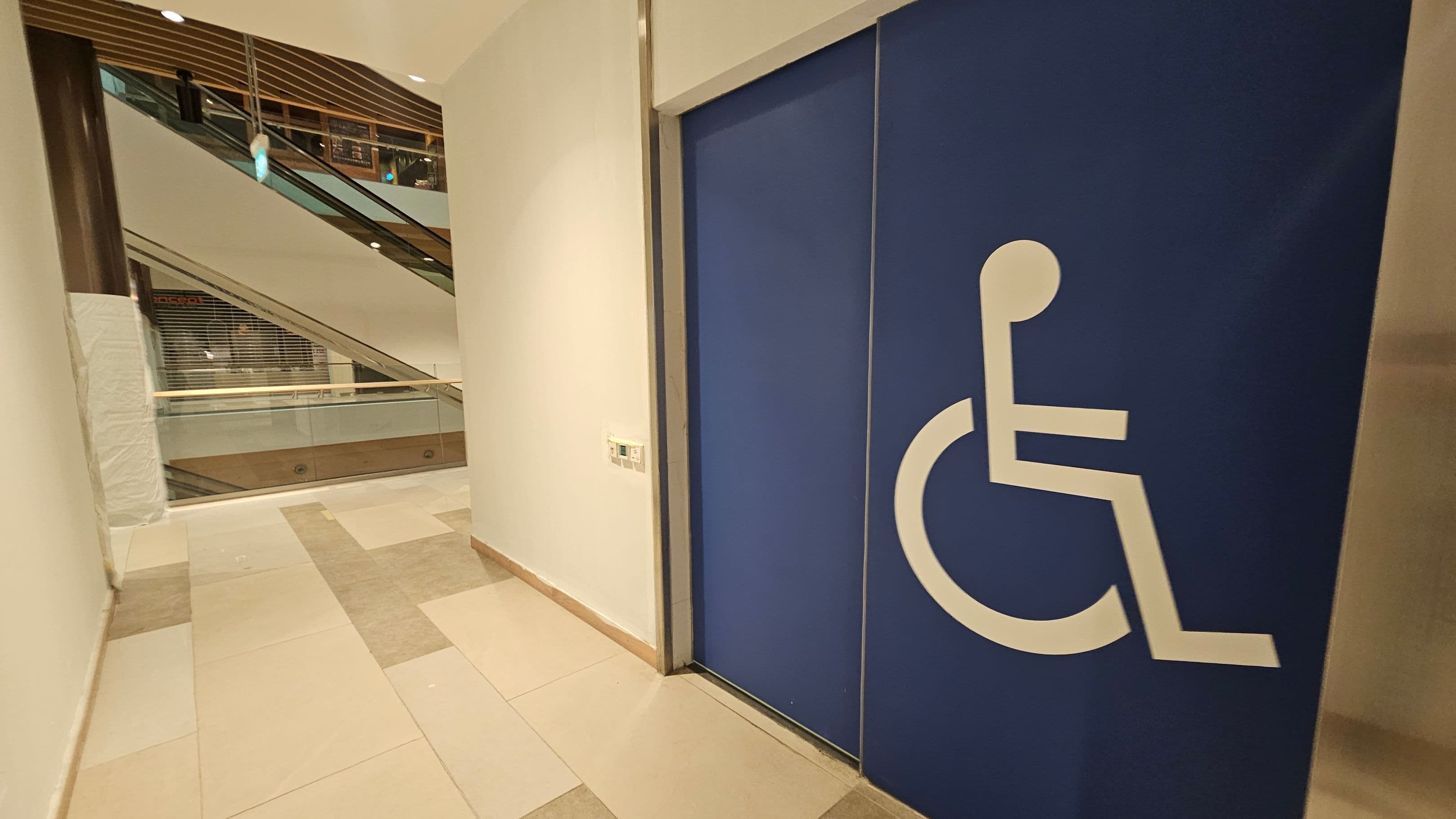 On the right, there is a large blue door with a white wheelchair symbol, indicating accessibility. The left side shows a corridor with a view of an escalator and glass railings. The floor is tiled in shades of beige and pink. The ceiling has recessed lighting and wooden slats.