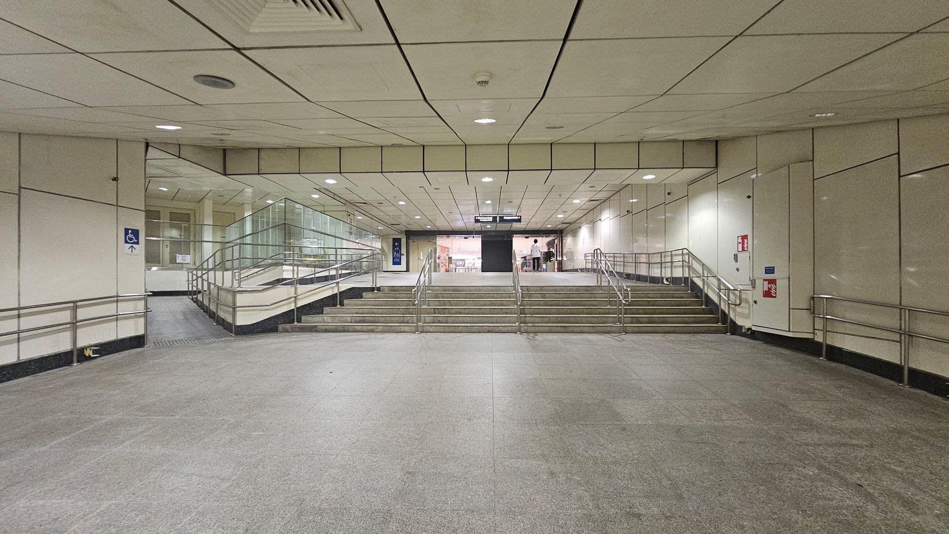 A spacious underground passage with a grey tiled floor and a light-coloured panelled ceiling. In the centre, a set of stairs with metal railings leads upwards, flanked by ramps on both sides for accessibility. The walls are lined with light panels, and there are signs indicating directions. At the top of the stairs, a glass entrance is visible, with a person standing near it. A fire extinguisher is mounted on the right wall.