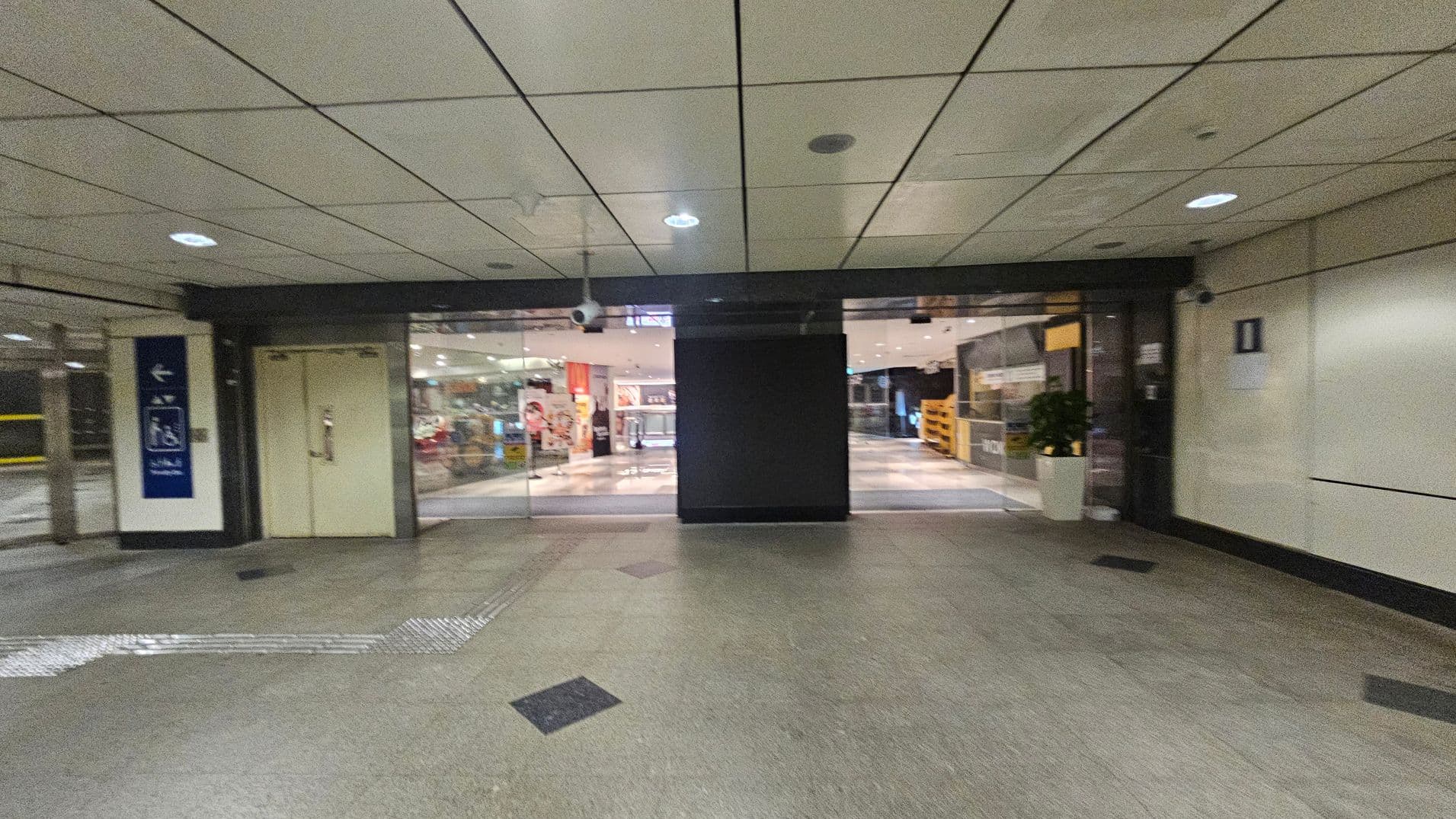 A corridor with a tiled ceiling and greenish floor. In the centre, there are glass doors leading to a brightly lit interior space with visible signage and displays. On the left, there's a sign with directional arrows and a closed door. On the right, a wall with a small sign and a potted plant near the entrance. The area is well-lit with ceiling lights.
