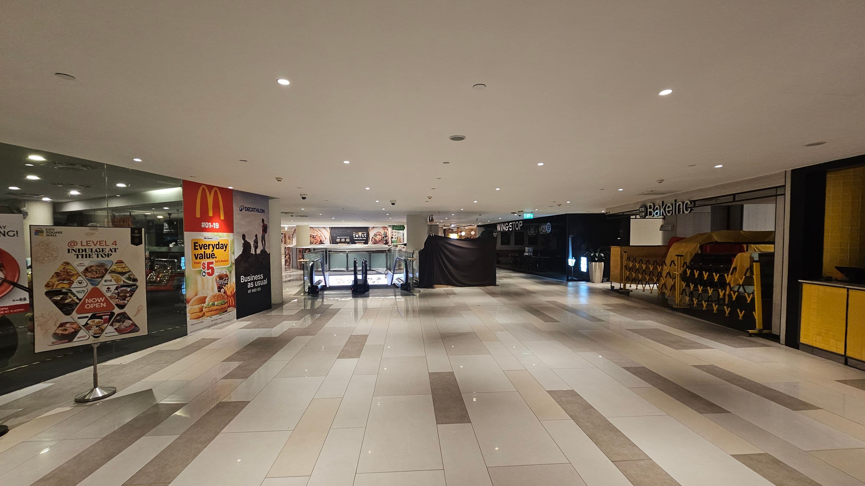 A shopping mall interior with a wide, tiled floor in shades of grey and white. On the left, there are promotional posters, including one for McDonald's. In the centre, an escalator is visible, leading to a lower level. On the right, there is a closed shop with a sign reading "Bake Inc" and a covered counter. The ceiling is white with recessed lighting, creating a bright atmosphere.