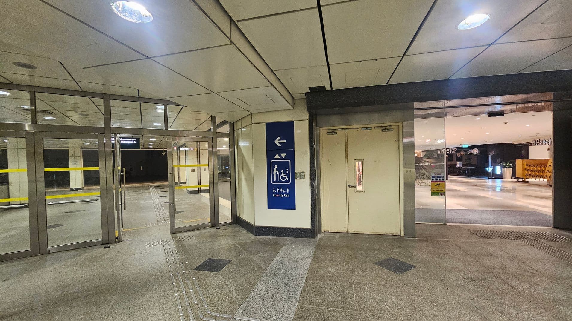 On the left, there are glass doors with yellow horizontal lines, leading to a corridor. In the centre, a blue sign indicates priority use for lifts, with symbols for accessibility. On the right, a closed door with a small window is adjacent to a glass entrance showing a glimpse of a brightly lit interior with shops. The floor is tiled with a mix of grey and black patterns, and the ceiling is white with recessed lighting.