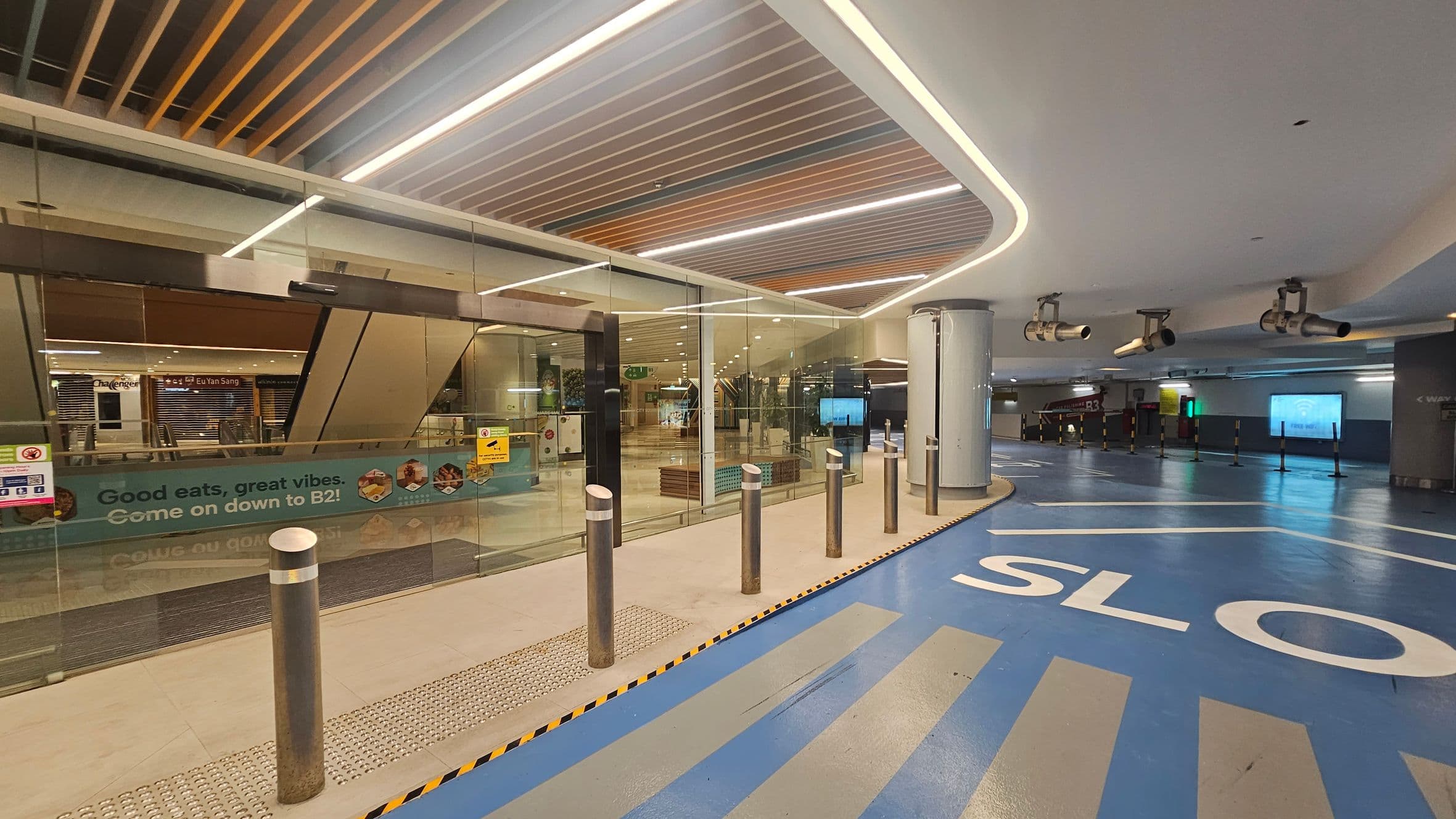 On the left, there is a glass entrance with a sign that reads "Good eats, great vibes. Come on down to B2!" Behind the glass, there are closed shopfronts, including one labelled "Eu Yan Sang." The floor is a mix of beige and blue, with the word "SLOW" painted in white. Metal bollards line the entrance. The ceiling features a modern design with recessed lighting and wooden slats. On the right, there are large cylindrical structures and overhead lights.