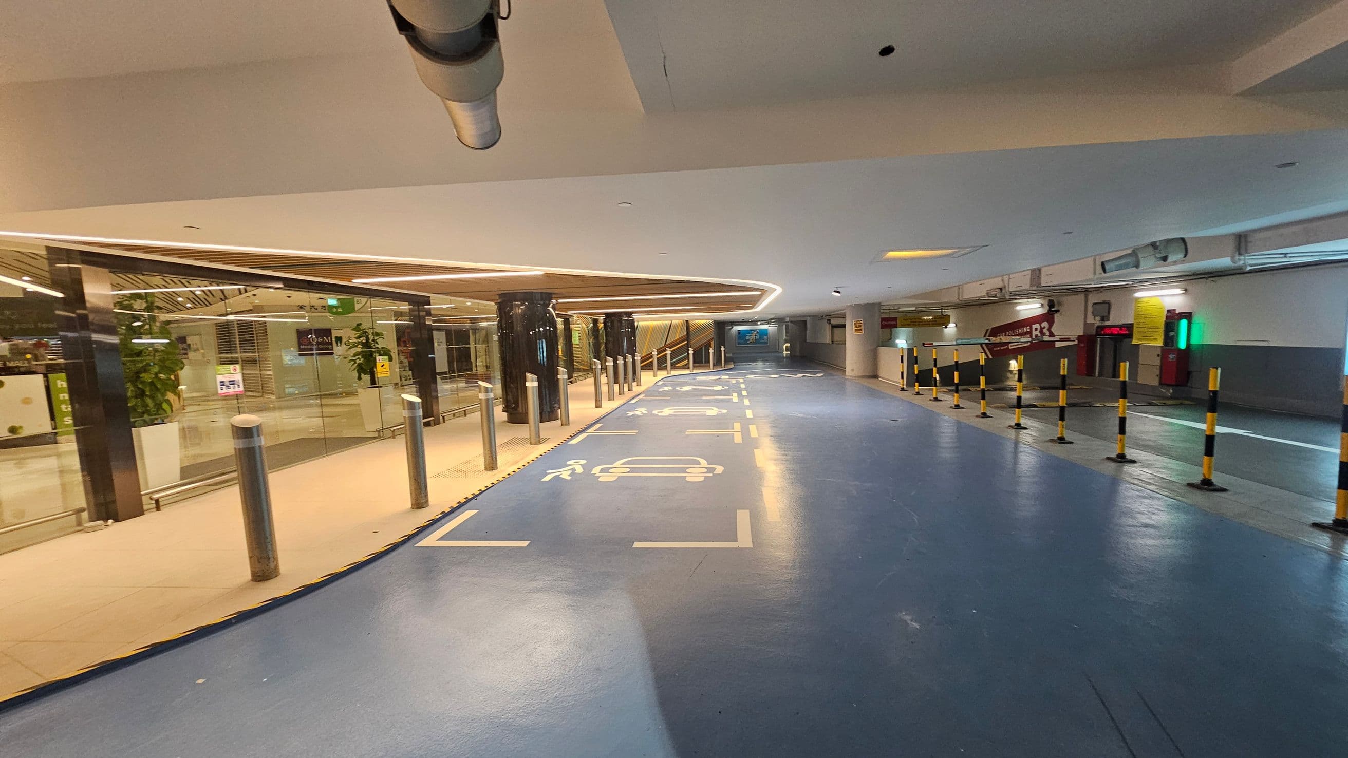 A parking area with a blue floor marked with white symbols for cars and pedestrians. On the left, there is a glass wall with plants and a view into a building interior. The ceiling has recessed lighting and a visible duct. On the right, there are yellow and black striped bollards and a sign indicating "R3". The background shows an escalator and more signage.