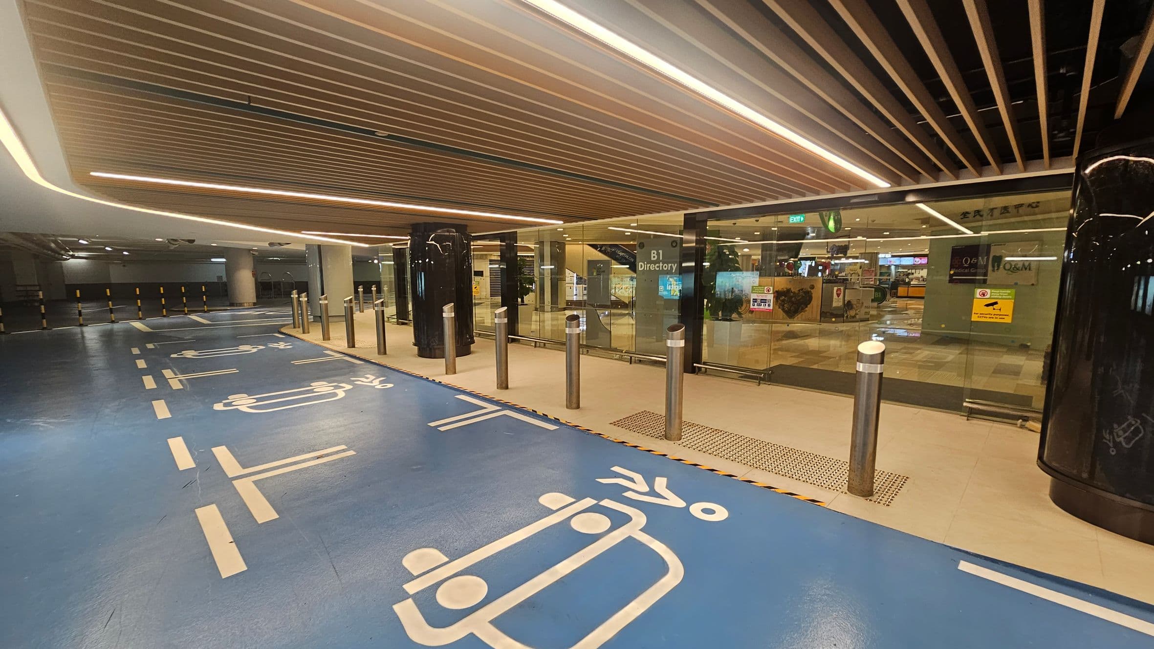 A modern underground parking area with a blue floor featuring white painted symbols for vehicles and pedestrians. On the right, there is a glass entrance with signs, including "B1 Directory," and several bollards in front. The ceiling has a wooden slat design with integrated lighting. In the background, there are visible shops and an exit sign. The left side shows more of the parking area with yellow and black striped barriers.