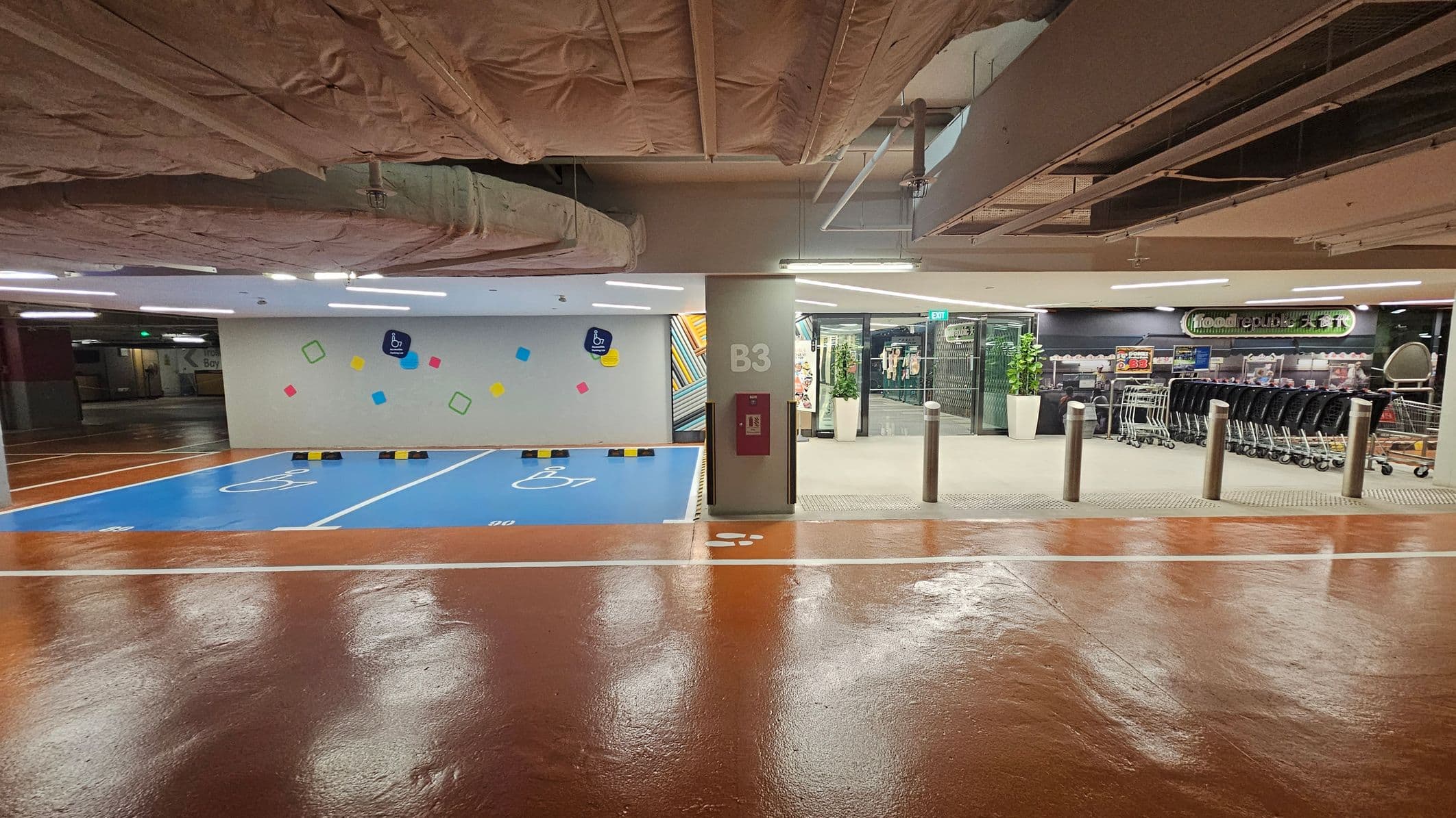 On the left, there are two blue parking spaces marked with wheelchair symbols, indicating accessibility. The wall behind is decorated with colourful geometric shapes. In the centre, a pillar marked "B3" has a fire hose cabinet attached. On the right, there is a supermarket entrance with shopping trolleys lined up outside. The floor is shiny and brown, and the ceiling has exposed ductwork and pipes.