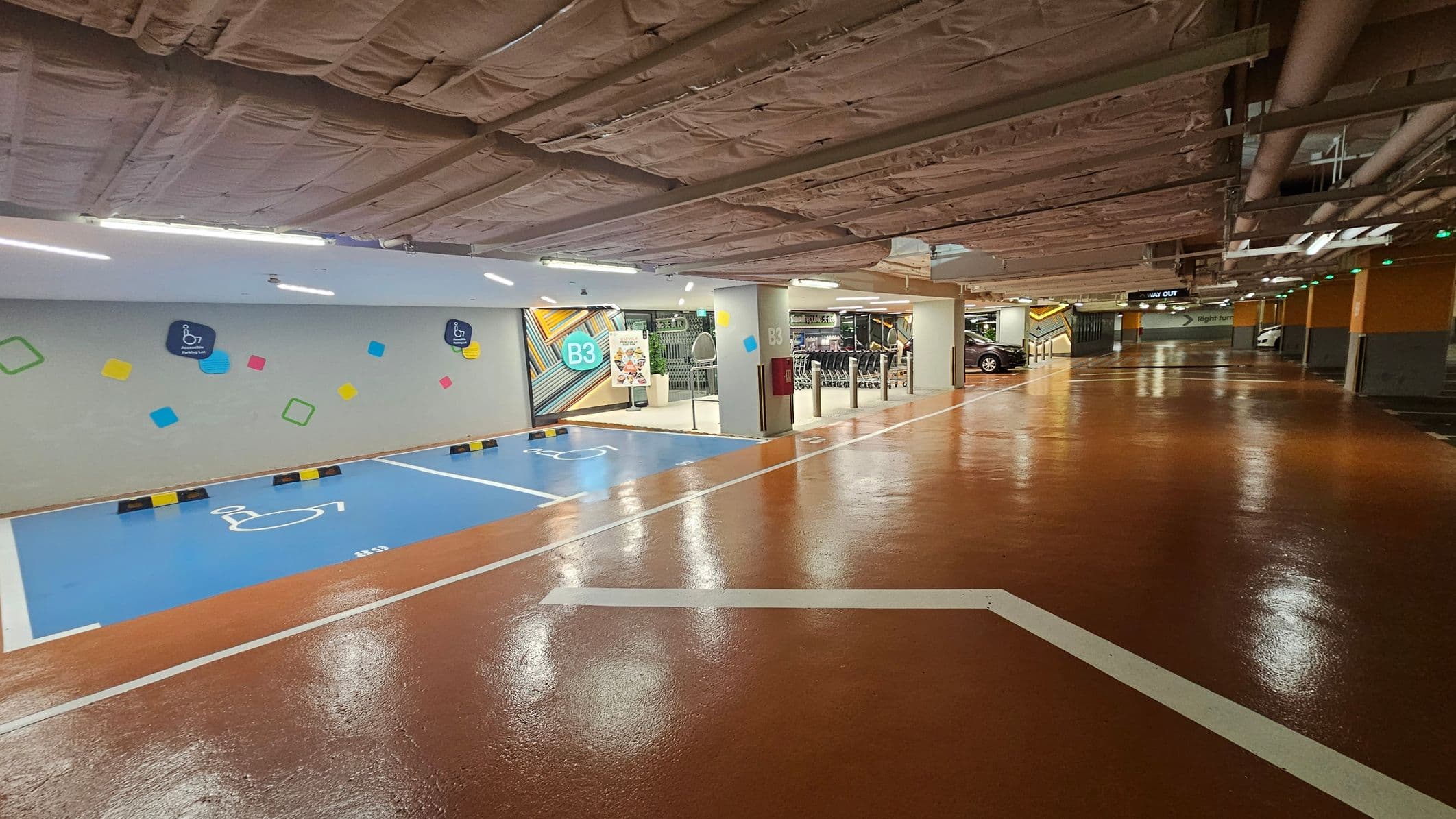 A parking garage with a shiny, brown floor and a ceiling covered in exposed insulation and pipes. On the left, there are blue parking spaces marked for disabled access, with colourful geometric shapes on the wall. In the middle, a sign indicates level B3, and there are shopping trolleys and a poster near the entrance. On the right, cars are parked, and a sign points to the way out. The area is well-lit with overhead lights.